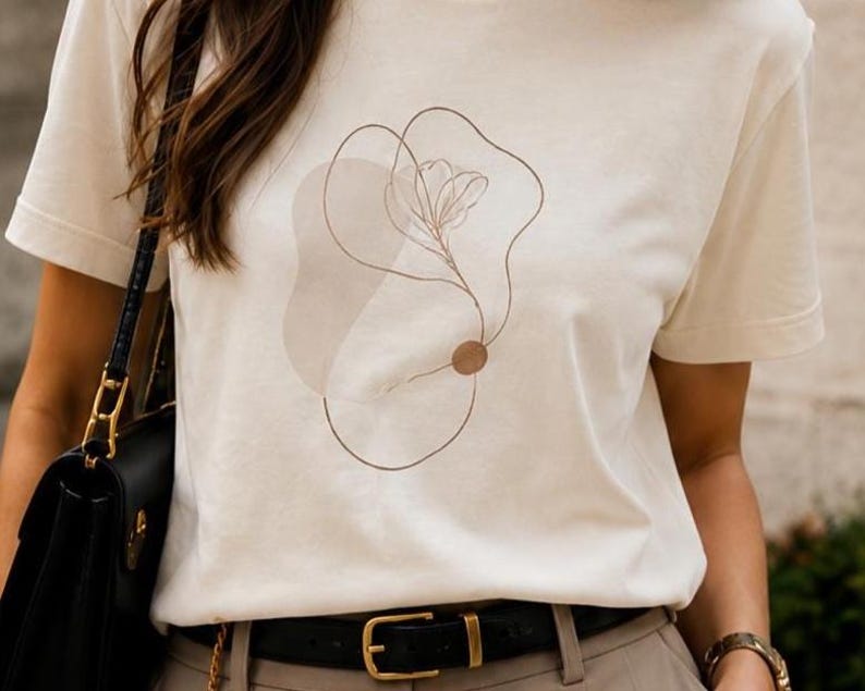 May include: A cream-colored short-sleeve t-shirt featuring a brown line art graphic of a flower and abstract shapes on the front. The design includes a thin, continuous line drawing of a floral stem and a solid brown circle accenting the composition.