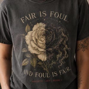 May include: A charcoal grey short-sleeve t-shirt featuring a graphic of a split rose, half white and half black. The text reads FAIR IS FOUL, AND FOUL IS FAIR. MACBETH ACT 1, SCENE 1 is printed in small red letters at the bottom of the graphic design.