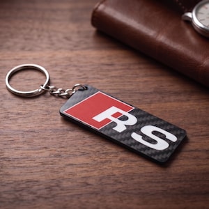 May include: A rectangular keyring featuring a carbon fibre pattern with a red square and the white letters RS printed on the surface. The keyring is attached to a silver metal split ring by a short chain, showcasing a sleek and modern design.