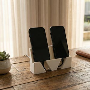 May include: A white phone charging station holds two black smartphones. The station is on a rustic wooden table, with a small succulent in a ceramic pot to the left. A notebook and a cup of coffee are also on the table.