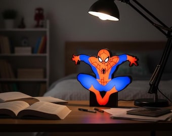 Könnte beinhalten: Nachtlicht in Form von Spider-Man in Blau und Orange. Die Lampe zeigt den Superhelden in einer dynamischen Pose. Sie steht auf einem Schreibtisch mit einer schwarzen Schreibtischlampe und offenen Büchern.