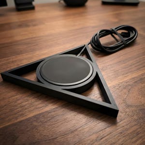 May include: A black wireless charger with a triangular frame and a circular charging pad. The charger has a black cable attached. The design is modern and minimalist, suitable for a desk or workspace.