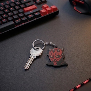 May include: A black and red keychain featuring a stylized image of Darth Maul's face. The keychain is attached to a silver key via a chain. The keychain is on a black surface, near a keyboard and mouse.