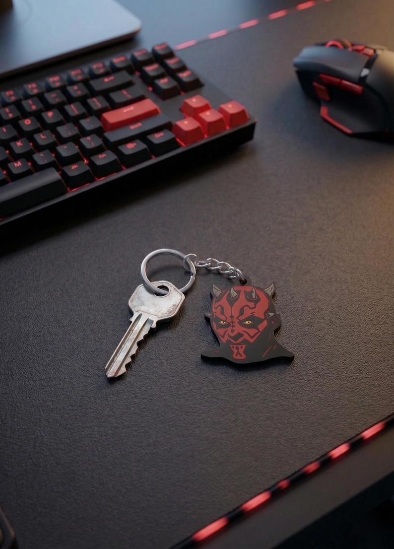 May include: A black and red keychain featuring a stylized image of Darth Maul's face. The keychain is attached to a silver key via a chain. The keychain is on a black surface, near a keyboard and mouse.