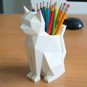 May include: A white, geometric cat-shaped pencil holder containing coloured pencils. The holder is made of a hard, smooth material and sits on a wooden desk. The pencils are various colours, including red, blue, green, and yellow.