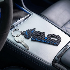 Puede incluir: Un llavero negro y azul con el texto "5.0 COBRA" se muestra sobre una superficie plateada junto a una llave de coche. El llavero tiene un patrón de fibra de carbono y un acento azul. El interior de un coche es visible en el fondo.