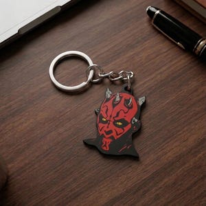 May include: A black and red keychain featuring the face of Darth Maul from Star Wars. The keychain has a silver ring and chain. The image is on a wooden surface with a laptop and pen in the background.