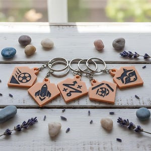 May include: A collection of keychains with peach-colored pendants and silver key rings. Each pendant features a different black graphic design. The keychains are arranged on a white wooden surface, surrounded by small stones and sprigs of lavender.