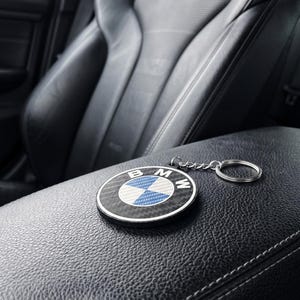 May include: A black and silver BMW keychain with the BMW logo, featuring a blue and white checkered design, rests on a black leather car armrest. The car seat is black leather with a textured surface.