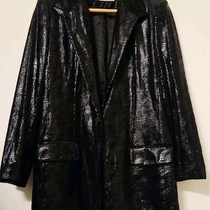 May include: A black blazer featuring a textured, reptilian-inspired pattern across the entire garment. The jacket includes a notched lapel, long sleeves, and two front flap pockets. It is displayed on a wooden hanger against a plain background.