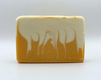 May include: A rectangular bar of handmade soap with a two-tone design. The bottom half is a vibrant yellow-orange, while the top half is a creamy white. Swirls of white cascade down the sides, creating a marbled effect. The soap is unscented.