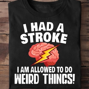 Puede incluir: Camiseta negra con el texto blanco "I HAD A STROKE I AM ALLOWED TO DO WEIRD THINGS!". El diseño incluye un cerebro rosa con un rayo amarillo.