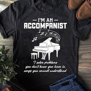 May include: A dark gray short-sleeve t-shirt featuring a white graphic of a grand piano with musical notes and the text "I'M AN ACCOMPANIST" followed by "I solve problems you don't know you have in ways you cannot understand" printed in white.