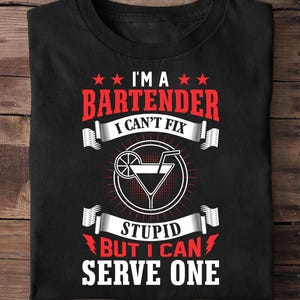 Könnte beinhalten: Ein schwarzes T-Shirt mit rotem und weißem Grafiktext, der I'M A BARTENDER I CAN'T FIX STUPID BUT I CAN SERVE ONE lautet. Das Design enthält ein zentrales weißes kreisförmiges Symbol eines Cocktailglases mit einem Strohhalm und einer Zitrusfruchtscheibe.