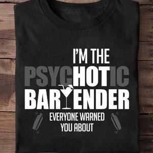 Pode incluir: Camiseta preta com texto branco: "I'M THE PSYCHOTIC BAR TENDER EVERYONE WARNED YOU ABOUT." Um copo de cocktail e duas coqueteleiras também estão presentes no design.