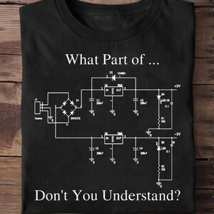 Pode incluir: Camiseta preta com um diagrama esquemático branco e o texto "What Part of... Don't You Understand?" O diagrama inclui componentes como uma ponte, resistores e capacitores.