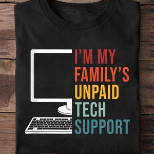 May include: Black t-shirt with a white computer monitor and keyboard graphic. Text reads "I'M MY FAMILY'S UNPAID TECH SUPPORT" in red, orange, yellow, teal, and blue. The shirt is folded on a wooden surface.