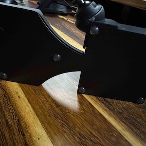May include: A black, angular mounting bracket featuring a spherical ball-joint connector at the top. The structure has a curved cutout section and includes several small, translucent circular adhesive bumpers attached to its flat surface.