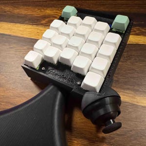 May include: A black custom mechanical keypad featuring a grid of white square keycaps, two light green accent keys at the top, and a black ergonomic palm rest with a ball-jointed joystick attachment. The base has a textured finish.