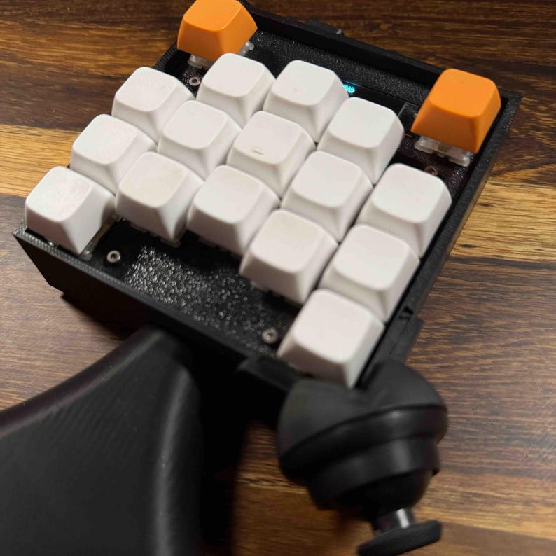 May include: A black macro keypad featuring a grid of twelve white square keycaps and two orange square keycaps at the top. The device has a textured black frame and a black joystick controller attached to the side, all mounted on a black base.