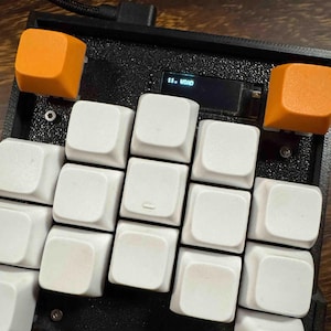May include: A black macro keyboard featuring a textured frame, twelve white square keycaps, and two orange square keycaps at the top corners. A small rectangular screen is positioned between the orange keys, displaying the text 11. USAD.