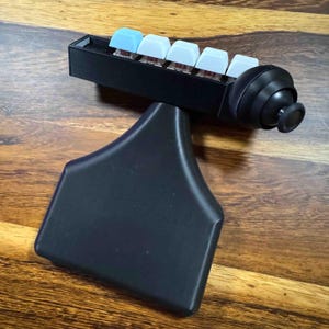 May include: A black handheld gaming controller featuring a row of five mechanical keyboard switches with white and light blue keycaps. The device includes a black joystick on the right side and a wide, flat, ergonomic handle base for grip.