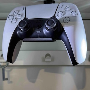 May include: A white and black video game controller hangs from a white ceiling mount. The controller has a white face with black buttons and a black handle.