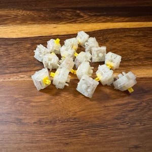 May include: A collection of clear, square mechanical keyboard switches with yellow stems, scattered on a wooden surface. The switches have a translucent appearance, revealing internal components. The background is a warm, brown wood grain.