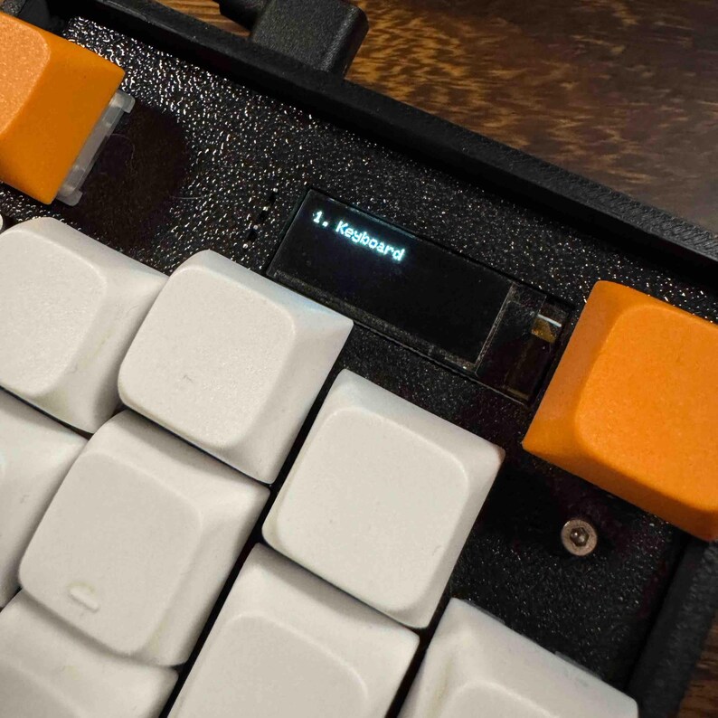 May include: A mechanical keyboard featuring a textured black frame, white keycaps, and orange accent keycaps. A small rectangular digital display screen is embedded in the frame, showing the white text 1. Keyboard on a black background.