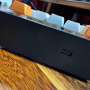 May include: A black mechanical keyboard case featuring a side-mounted USB-C port. The keyboard is equipped with a row of white keycaps and orange keycaps at the ends, showcasing a clean, functional design with a visible port for connectivity.