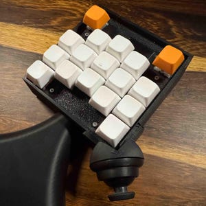 May include: A black mechanical keypad featuring a grid of twelve white keycaps and two orange keycaps. The device includes a black palm rest and a black ball-and-socket joint mount attached to the base, providing an adjustable ergonomic setup.