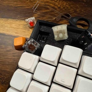 May include: A mechanical keyboard with white keycaps, a black foam base, and several loose components including a single orange keycap, a red switch, a brown switch, a white switch, a black switch, and a metal wire keycap puller tool.