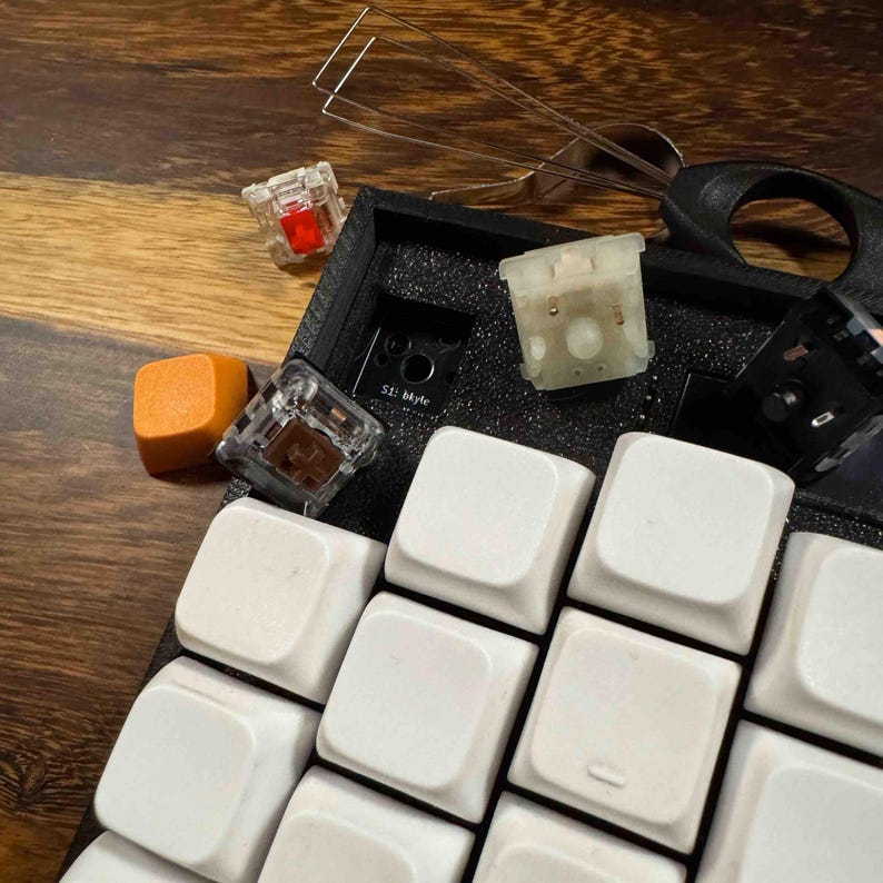 May include: A mechanical keyboard with white keycaps, a black foam base, and several loose components including a single orange keycap, a red switch, a brown switch, a white switch, a black switch, and a metal wire keycap puller tool.