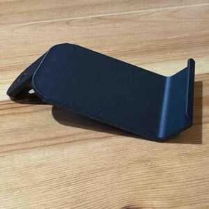 May include: A black plastic phone stand with a curved design. The stand is designed to hold a phone upright.