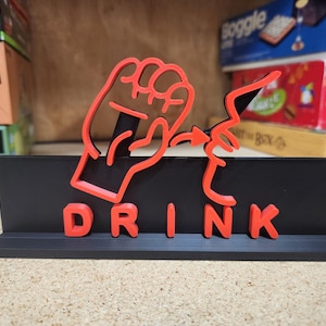 May include: A black tabletop sign with the word "DRINK" in red, stylized letters. Above the word is a red outline of a hand gesturing towards a lightning bolt. The sign is a decorative item.