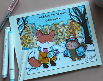 May include: A coloring book titled "Ash & Echo: The Big Apple Coloring Book" featuring a cartoon fox and owl in a snowy city scene. The image includes white Ohuhu art markers and a pink pen, all on a light brown surface.