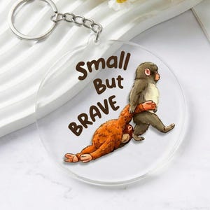 May include: A clear, round keychain with a silver keyring. The keychain features a cartoon illustration of two monkeys, one brown and one orange, with the text "Small But Brave" in brown lettering.
