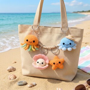 May include: A beige tote bag with four crochet keychains: an orange octopus, a blue octopus with a hat, a pink clam with a pearl, and an orange starfish. The keychains are hanging from the bag's metal rings. The background is a sandy beach and ocean.