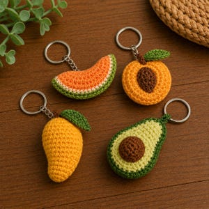 May include: Four crochet fruit keychains: a watermelon slice, a peach, a mango, and an avocado. Each keychain features a silver ring and chain. The watermelon is orange with a green rind, the peach is orange with a brown pit, the mango is yellow with a green leaf, and the avocado is green with a brown pit.