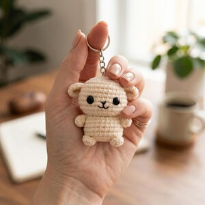 May include: A beige crocheted lamb keychain with black eyes and a smiling mouth. The lamb has small ears and arms. The keychain is held by a silver ring. The background is blurred, showing a desk and a cup of coffee.