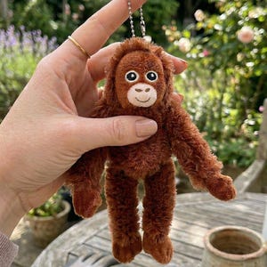 May include: A small, plush orangutan keychain in shades of brown. The toy has a detailed face with large eyes and a smiling mouth. It is held up by a silver chain, suggesting it can be attached to keys or a bag. The background is blurred, with greenery.