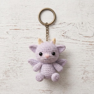 May include: A lavender crochet keychain featuring a small, adorable dragon with black eyes, small wings, and tan horns. The dragon is attached to a metal keyring and chain. The handmade plushie is sitting on a white wooden surface.