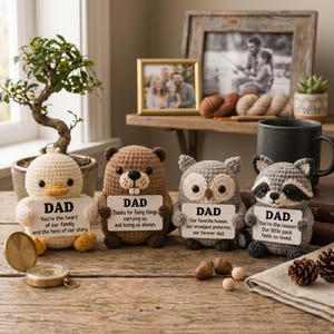 May include: Four handmade crochet animal figurines: a duck, a beaver, an owl, and a raccoon. Each holds a sign with "DAD" and a message. The duck says, "You're the heart of our family." The beaver says, "Thanks for fixing things." The owl says, "Our favorite human." The raccoon says, "You're the reason."