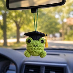 May include: A crocheted green frog car charm wearing a black graduation cap with a yellow tassel. The frog has large black eyes and is hanging from a green string, suspended from a car's rearview mirror. The background is blurred with green trees.
