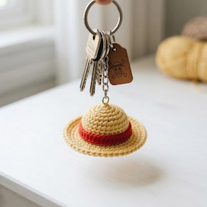 May include: A keychain with keys and a crochet hat charm. The hat is tan with a red band. A brown leather tag with a skull and crossbones is also attached to the keychain.