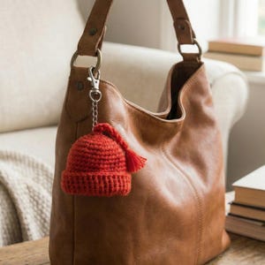 May include: A small, crocheted red beanie with a tassel charm is attached to a brown leather handbag via a silver-colored chain and clasp. The bag is a warm, medium brown color and has a shoulder strap. The background includes a cream-colored sofa and a stack of books.