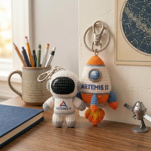 May include: Two crochet keychains: an astronaut and a rocket. The astronaut is white with a black visor and the text "ARTEMIS II". The rocket is white with orange and blue accents and the text "ARTEMIS II".