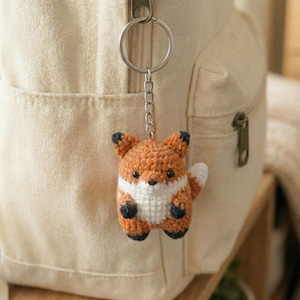 May include: A crocheted fox keychain in orange, white, and black hangs from a silver keyring. The fox has a white chest and belly, black paws and ears, and a white-tipped tail. The keychain is attached to a beige backpack.