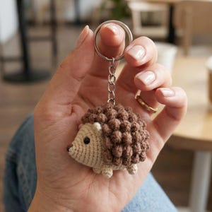 May include: A handmade crochet hedgehog keychain in shades of brown and cream. The keychain features a small, detailed hedgehog with a textured body and a metal keyring. The item is held in a person's hand.