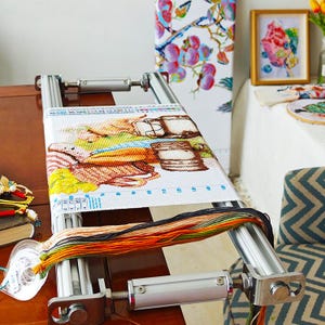 Puede incluir: Un marco de punto de cruz plateado con un diseño de punto de cruz colorido de una escena de dibujos animados. El marco tiene una perilla en el lateral para ajustar la tensión de la tela. El marco está sobre una mesa de madera con una pila de hilo de bordar de colores.